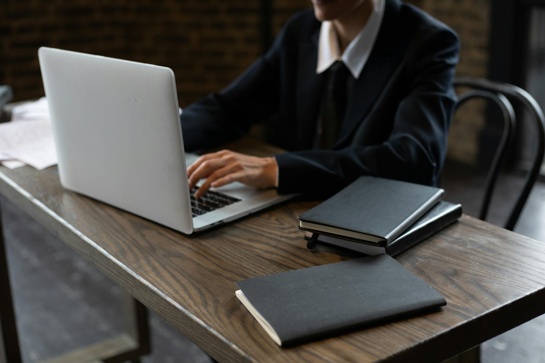 Close-up of a person in a suit typing on a laptop at a wooden desk with notebooks.
